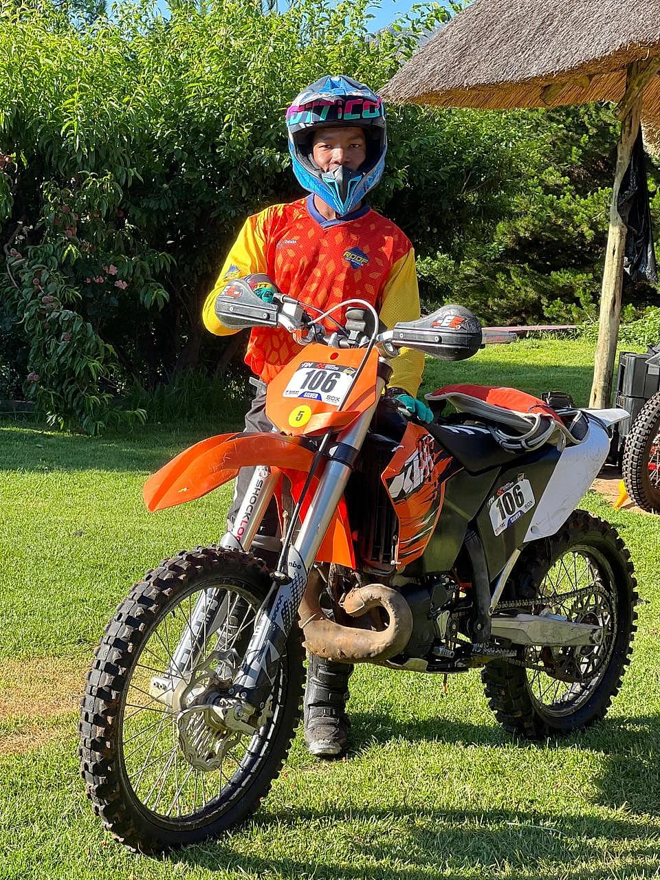 Helmeted rider sitting on an orange enduro motorcycle outside a thatched lodge
