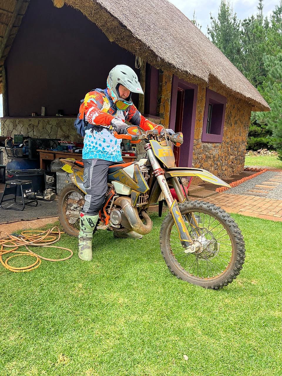 Helmeted rider preparing an enduro motorcycle beside a thatched lodge